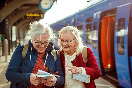 Zwei ältere Frauen stehen am Bahnhof und schauen lachend auf eine Karte. Im Hintergrund ist ein Zug am Gleis.
