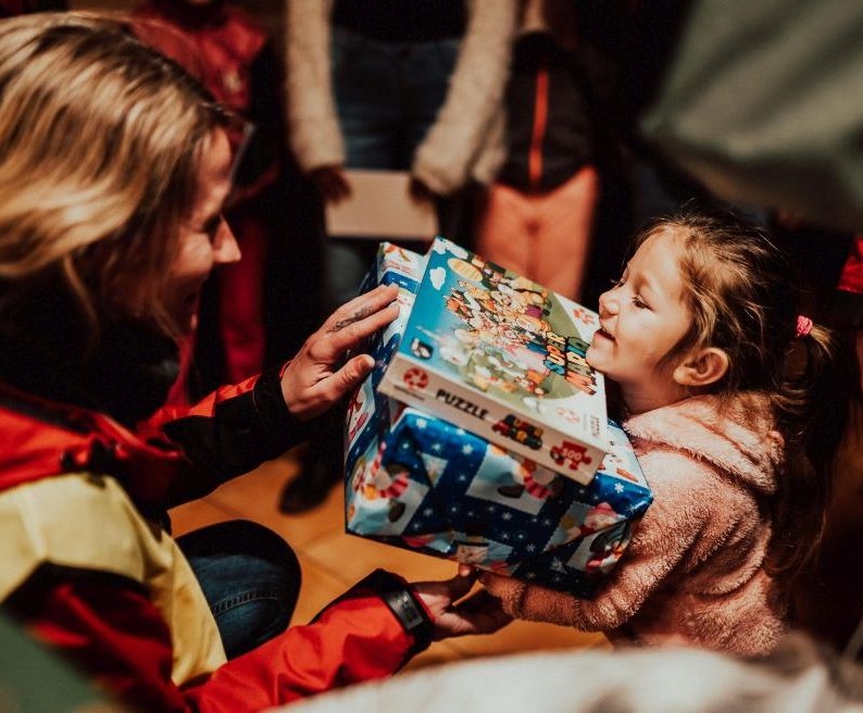 Eine Frau gibt einem kleinen Mädchen zwei große Geschenke. Beide lachen sich gegenseitig an und im Hintergrund sind mehr Kinder zu sehen.
