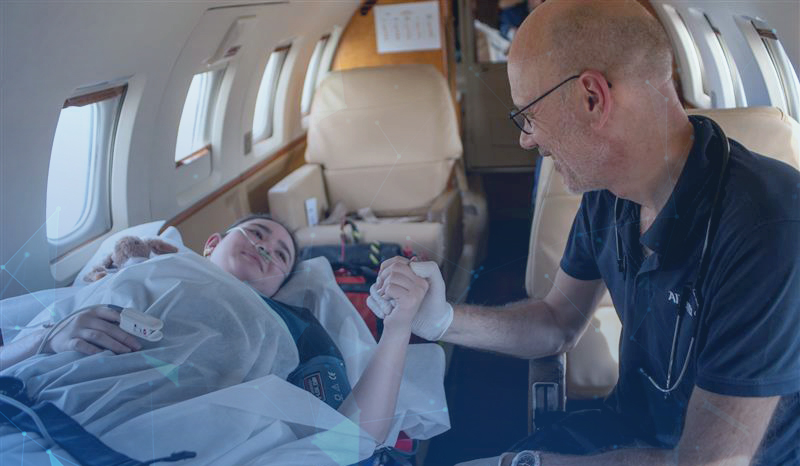 Eine Frau (Patientin) liegt mit Sauerstoffbrille und Pulsmesser am Finger auf einer Liege in einem Flugzeug. Neben ihr sitzt ein Mann (Arzt), um dessen Hals ein Stethoskop hängt. Er hält die Hand der Frau und trägt selbst einen weißen Handschuh.